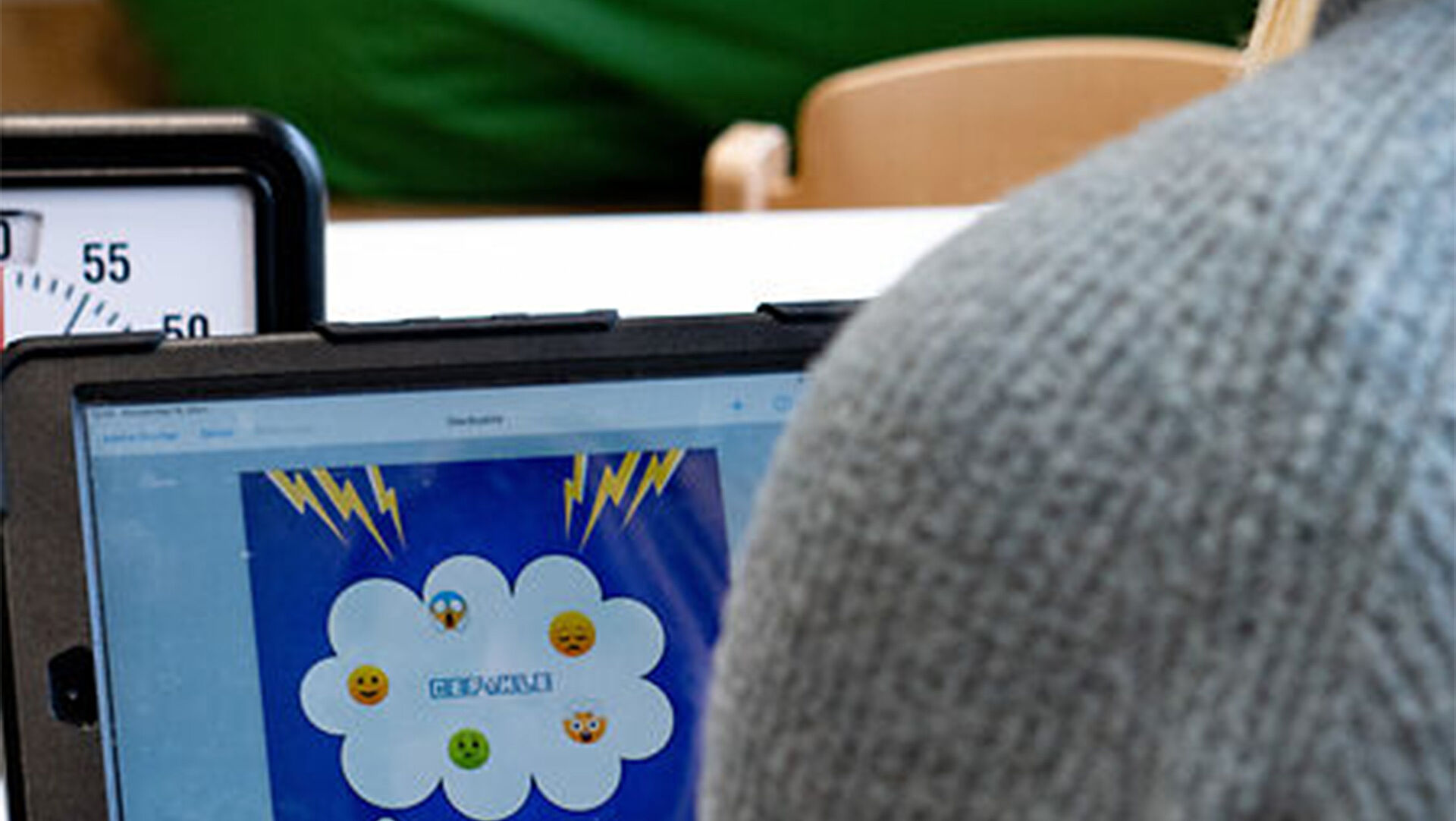 Begleitung_Startseite Im Vordergrund sieht man die linke Schulter eines Menschen, der vor einem Tablet sitzt. Auf dem Tablet des Bildschirms ist eine Grafik, auf der Gefühle mit Hifle von Emojis dargestellt sind.